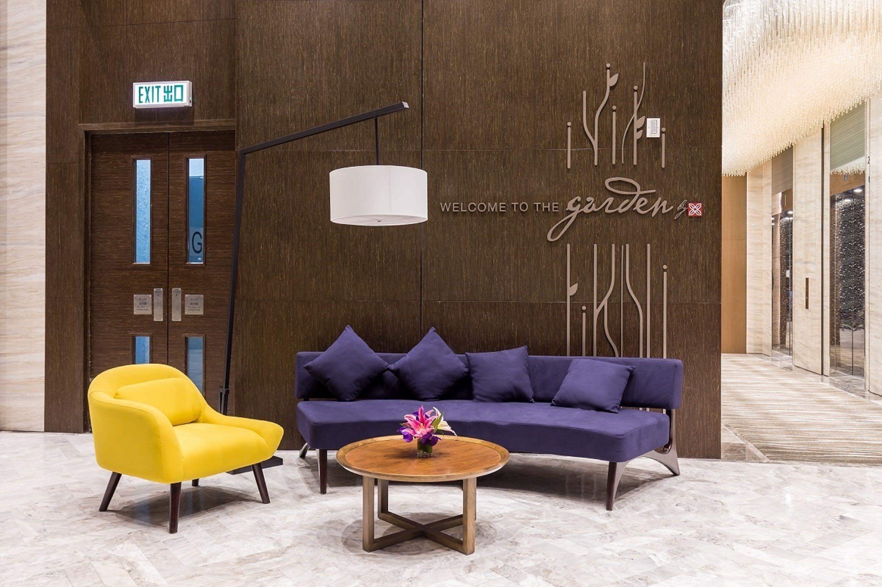 Un hall con un sofá púrpura, un sillón amarillo y una mesa de centro redonda de madera con un centro de flores.