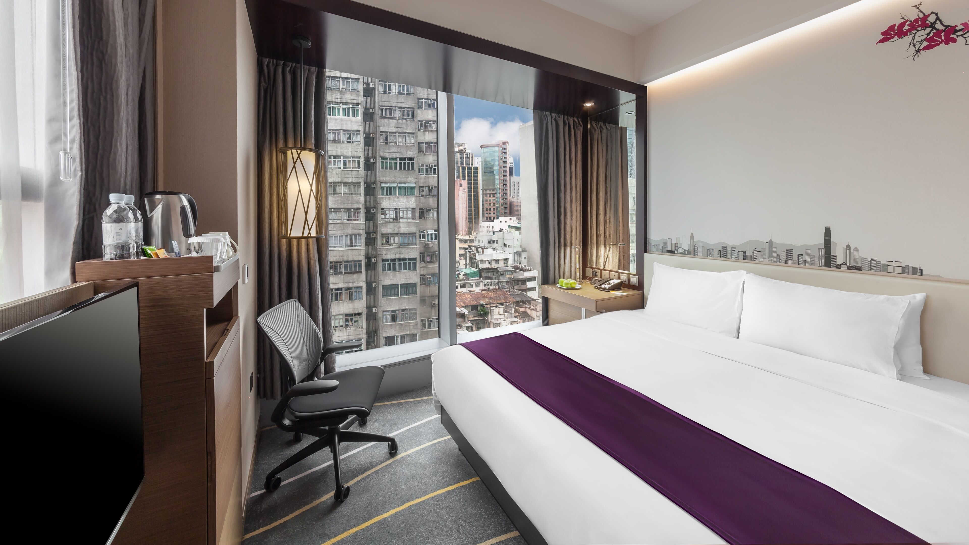 A hotel room with a large bed, a desk with a chair, a TV, and a view of a cityscape through a large window.