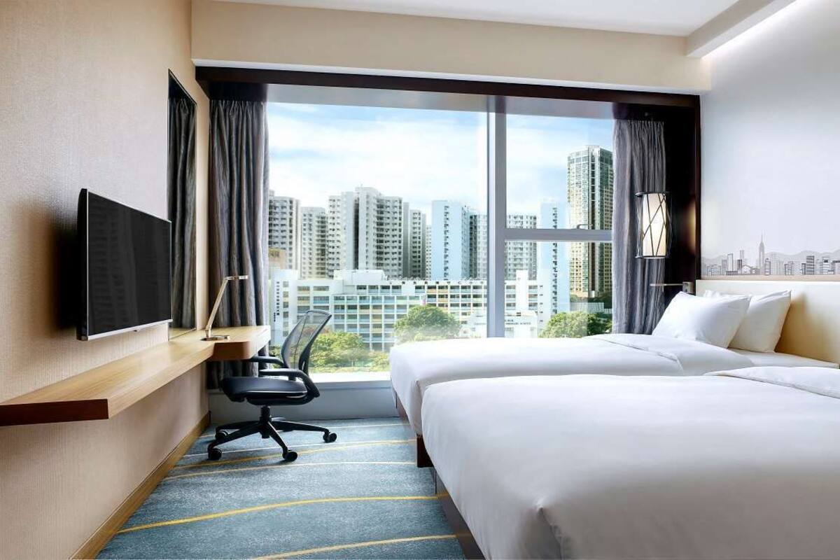 A hotel room with two beds, a desk, a chair, a TV, and a large window with a city view.