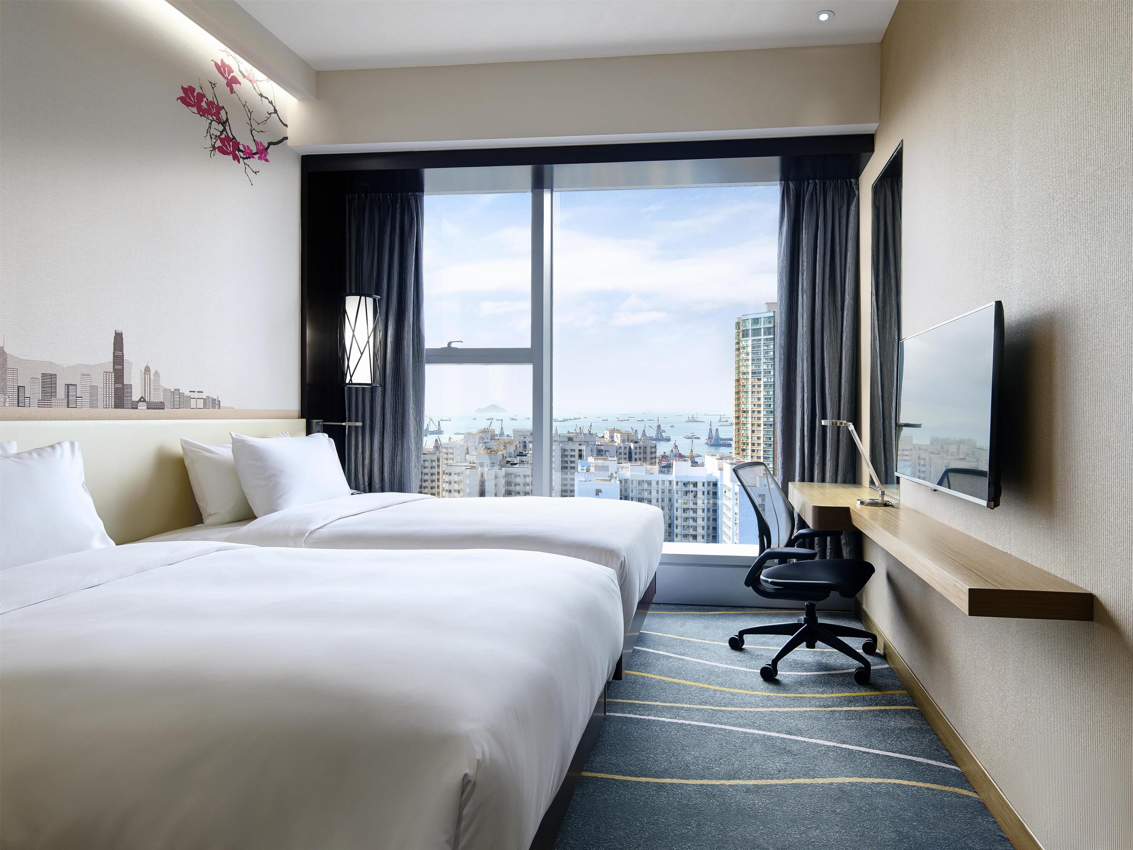 Een hotelkamer met twee bedden, een bureau, een stoel, een televisie en een groot raam met uitzicht op de stad.