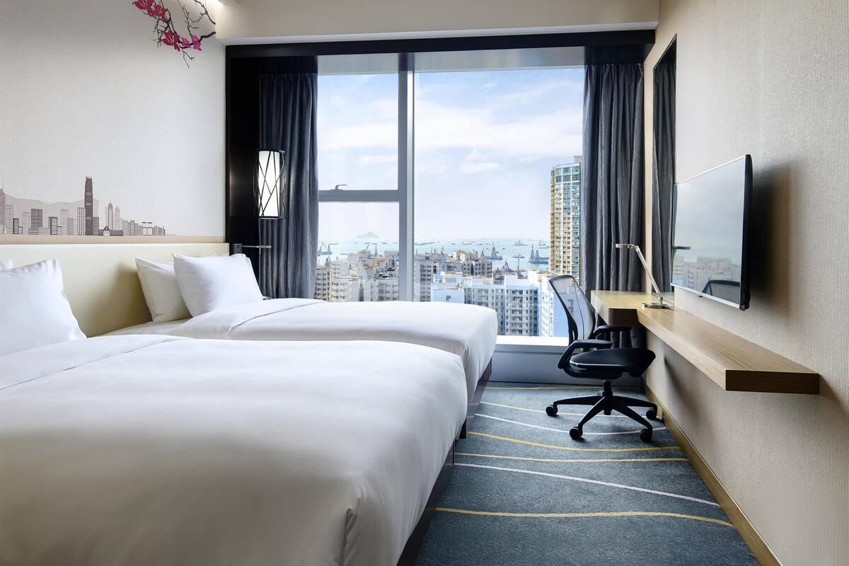 A hotel room with two beds, a desk, a chair, a TV, and a large window with a city view.