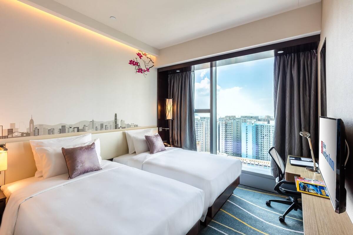 A hotel room with two beds, a desk, a chair, a TV, and a large window with a cityscape view.