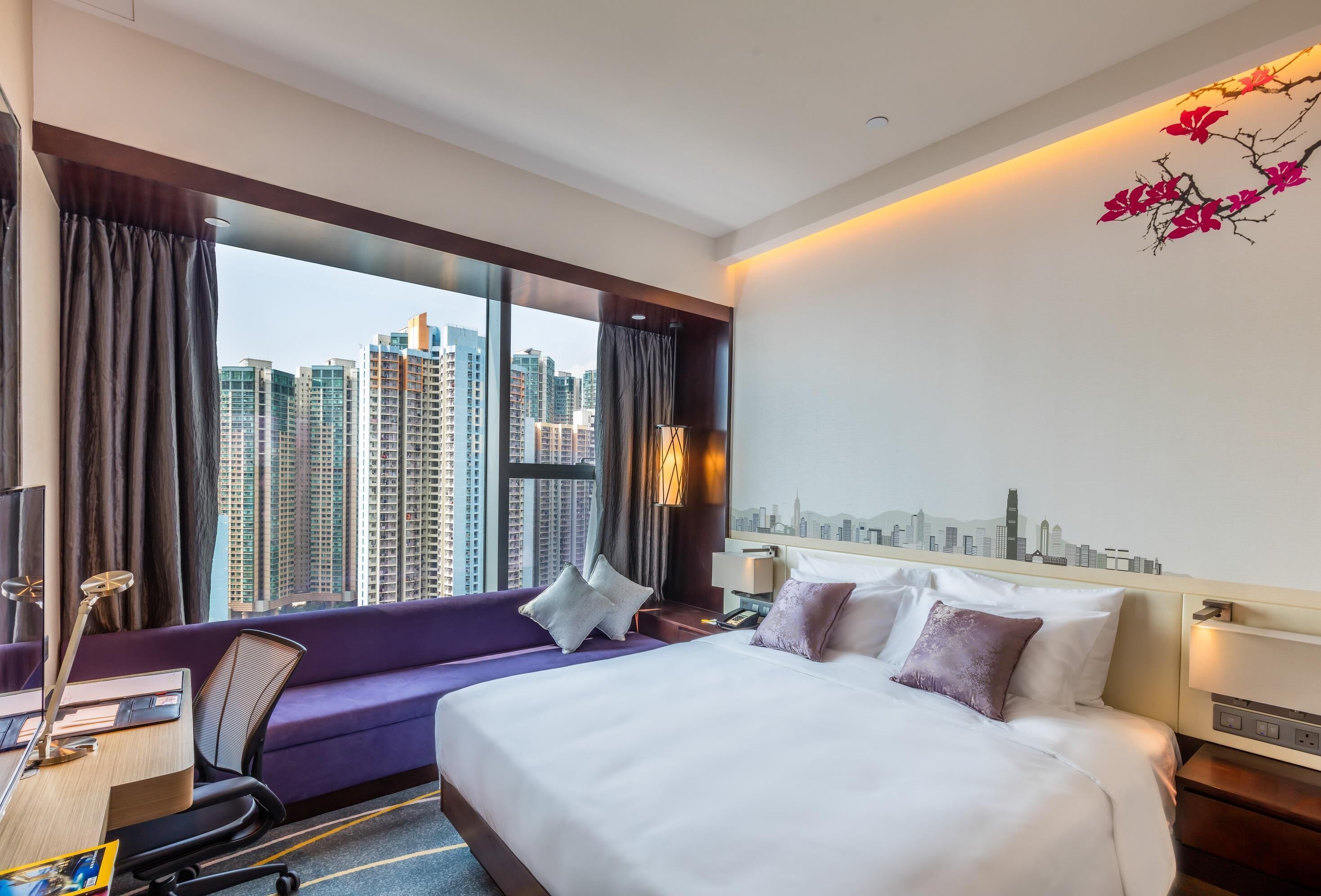 Hotelkamer met een bed, een bureau, een stoel en uitzicht op hoge gebouwen door grote ramen.