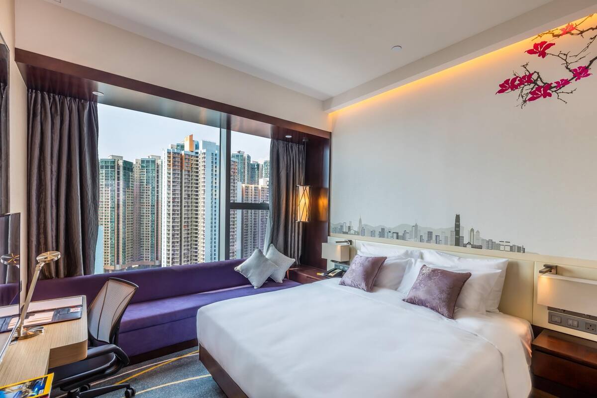 A hotel room with a bed, a desk, a chair, and a view of tall buildings through large windows.