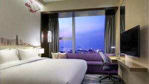 A hotel room with a large bed, a desk with a chair, a TV, and a window with a cityscape view.