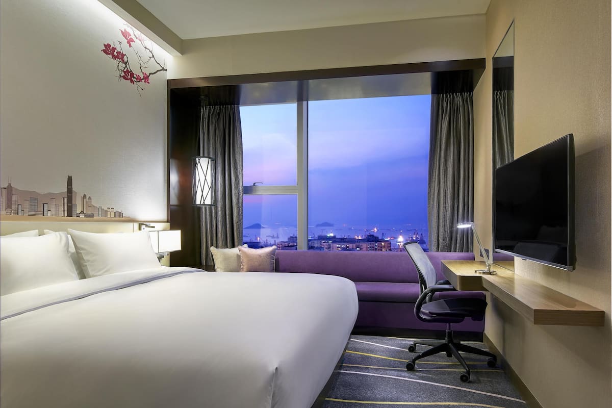 A hotel room with a large bed, a desk with a chair, a TV, and a window with a cityscape view.