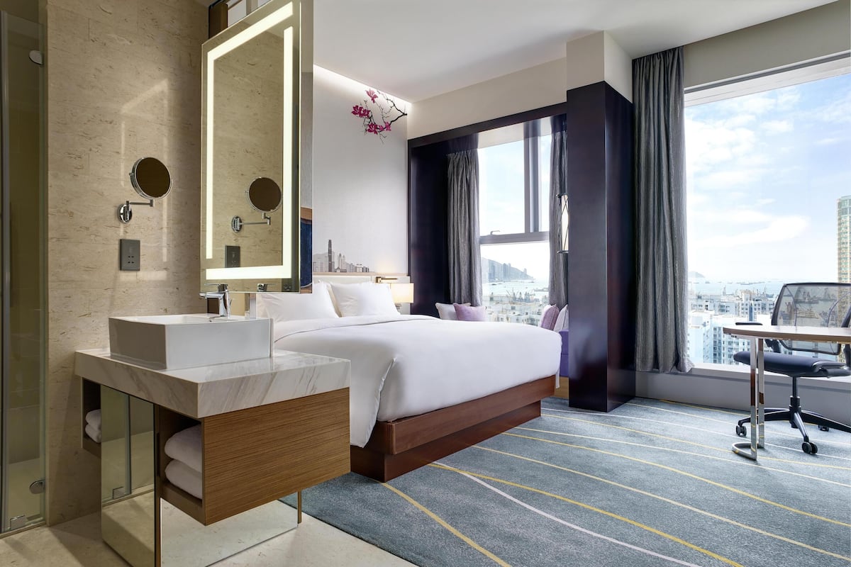 A modern hotel room with a large bed, a bathroom with a vanity, a large window with a city view, and a desk with a chair.