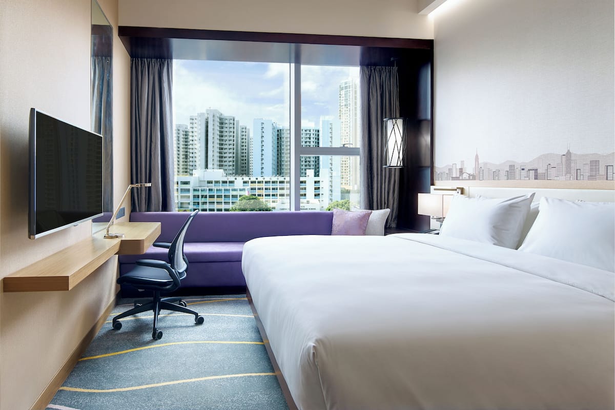 A hotel room with a bed, a desk, a chair, a TV, and a large window with a city view.
