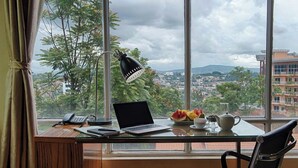 A room with a desk, laptop, telephone, and a view of a cityscape through large windows.