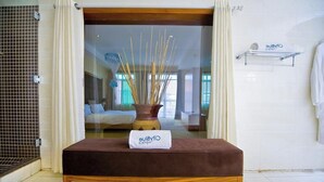 A hotel room with a bed, a bedside table with a towel, a shower area, and a view of the room through a glass door.