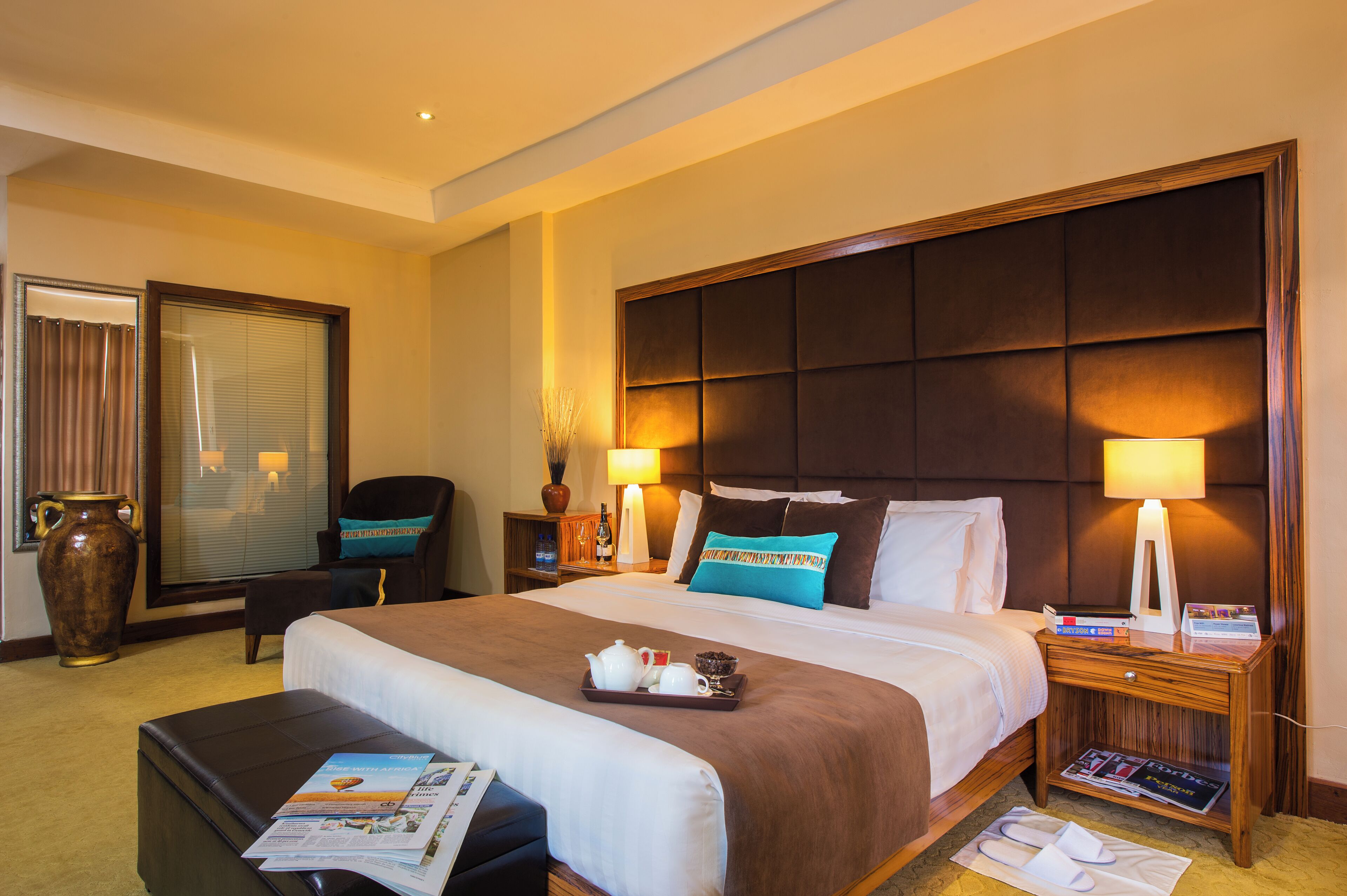 A hotel room with a large bed, a nightstand with a lamp, a chair, a large vase, and a window with curtains.