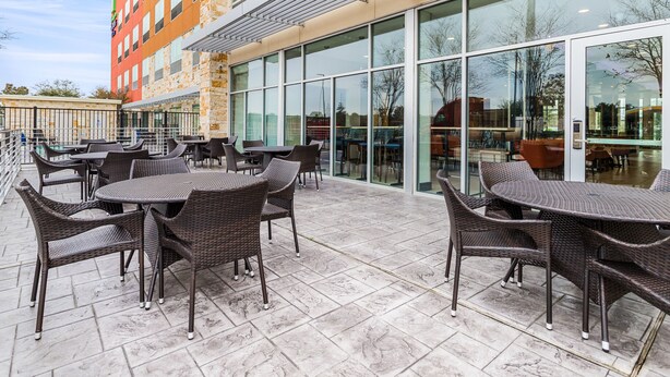 An outdoor patio area with wicker furniture and a stone-tiled floor, adjacent to a building with large glass windows. - Holiday Inn Express & Suites Houston NW - Cypress Grand Pky by IHG (Cypress)