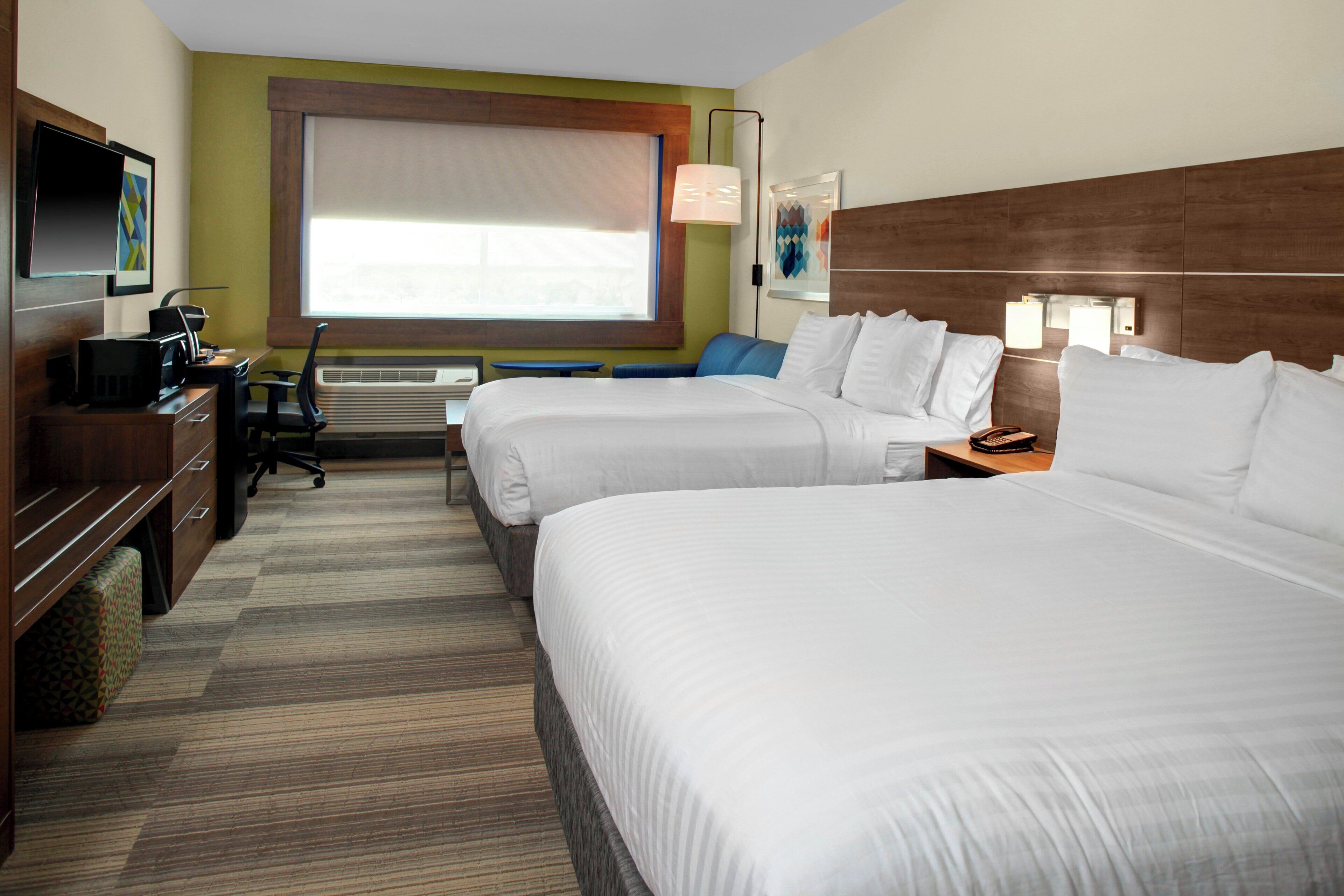 A hotel room with two beds, a desk with a computer, a chair, an air conditioning unit, and a television on the wall.