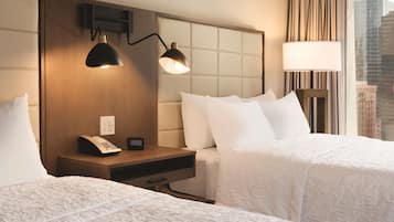 Habitación de hotel con dos camas, una mesita de noche con teléfono y reloj digital, una lámpara de pared y vista a la ciudad.