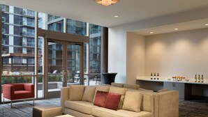 A modern hotel lobby with a beige sofa, a red armchair, a coffee table, and a kitchen area with wine bottles and glasses. - Hampton Inn Chicago West Loop Fulton Market Area (Chicago)