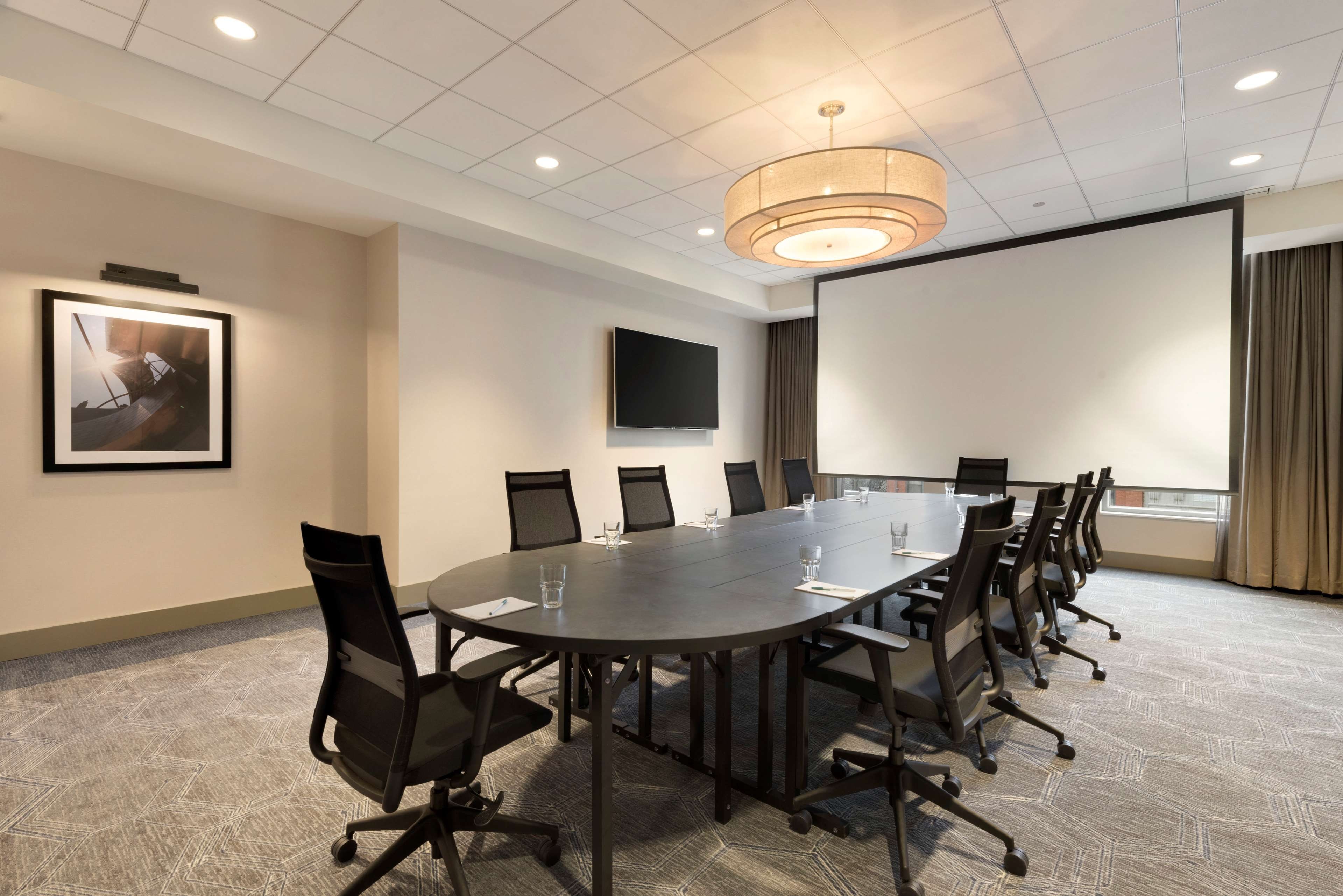 A conference room with a long table, chairs, a projector screen, and a television on the wall.