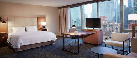 Habitación de hotel con cama, escritorio, silla, televisión y ventanas amplias con vista a la ciudad.