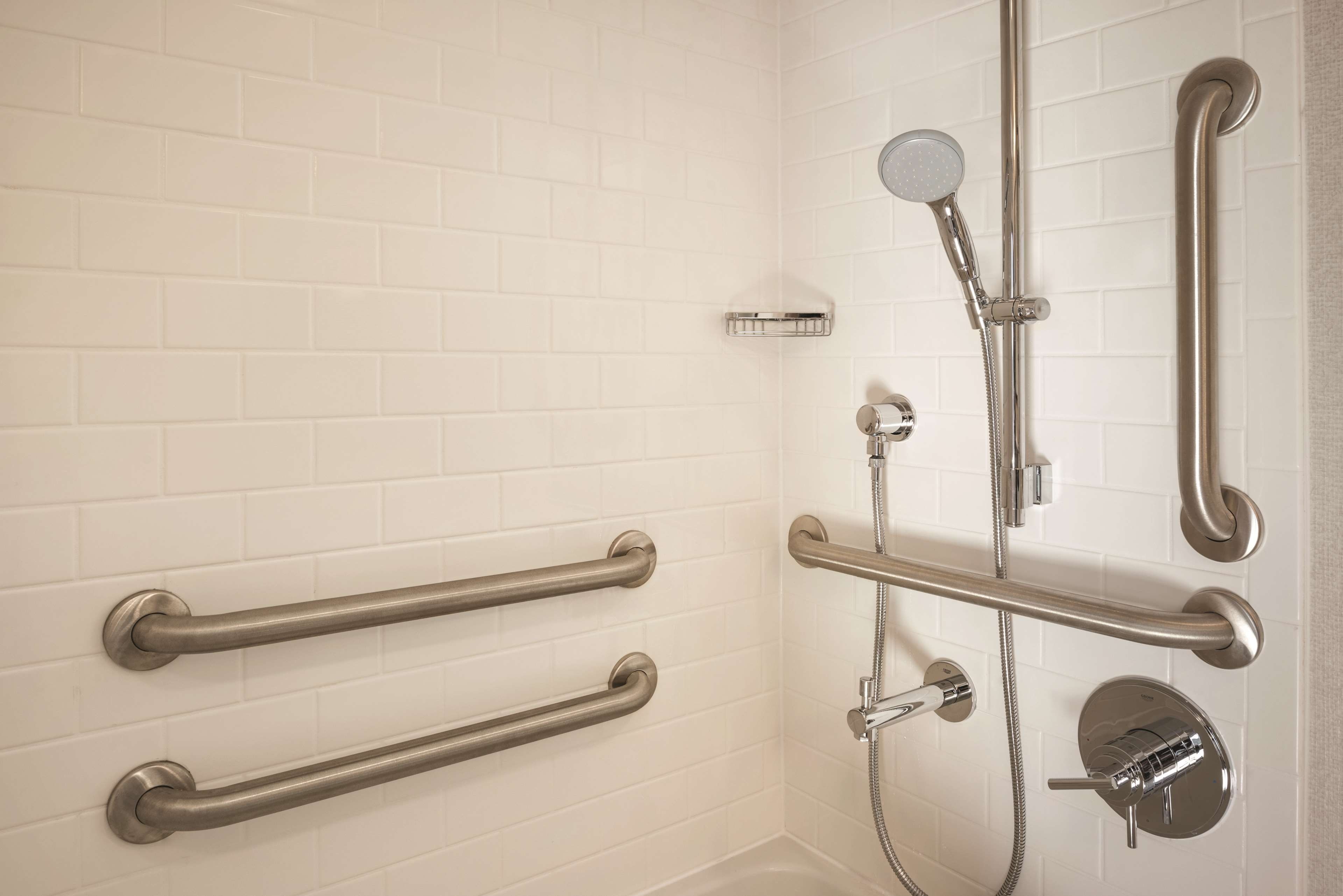 Une salle de bain avec des murs carrelés de carreaux blancs, une douche équipée d’un pommeau de douche à main et de deux barres d’appui fixées au mur.