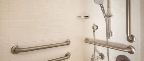 Une salle de bain avec des murs carrelés de carreaux blancs, une douche équipée d’un pommeau de douche à main et de deux barres d’appui fixées au mur.