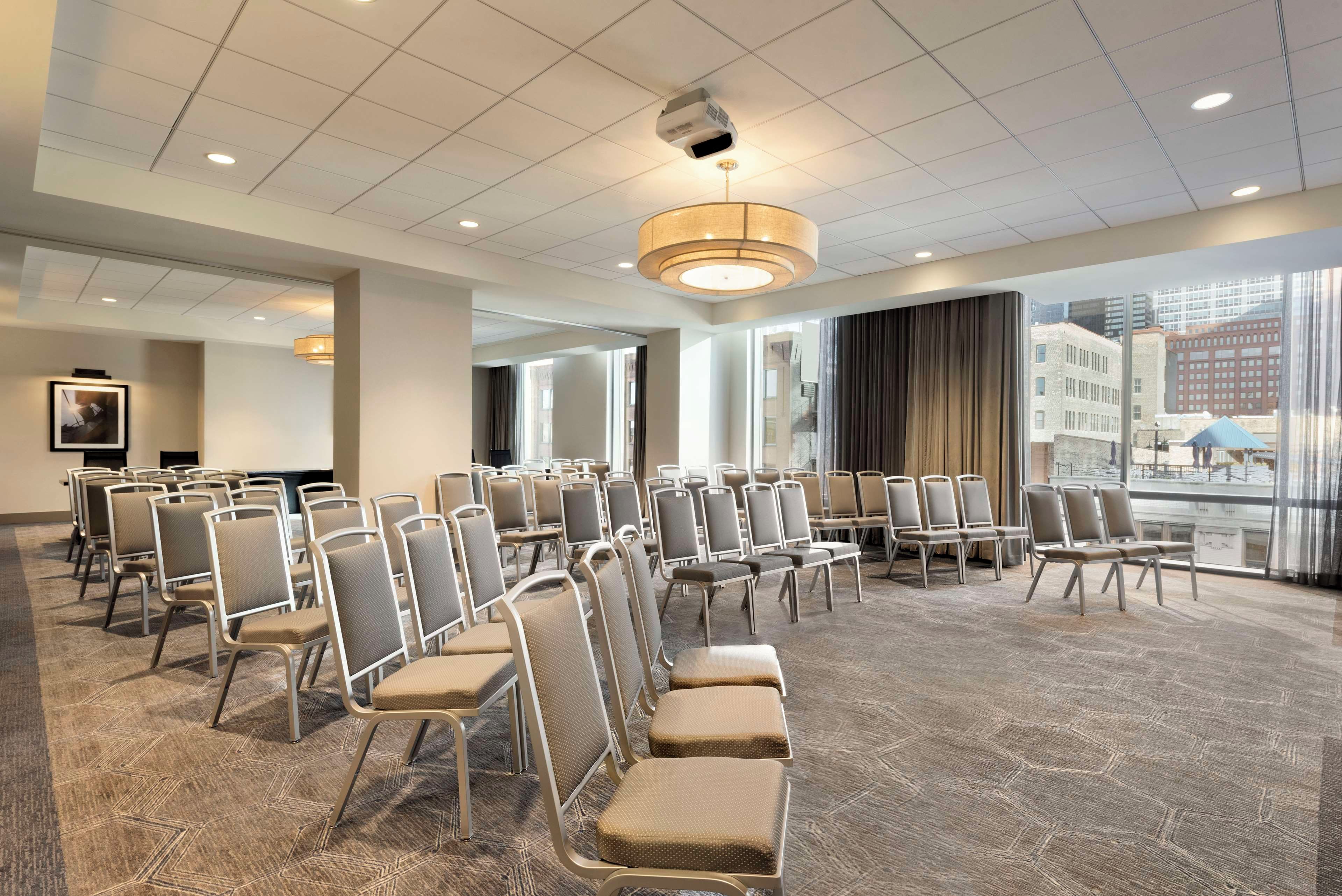 Un salón de conferencias con filas de sillas, un ventanal con vistas a edificios e iluminación empotrada en el techo.