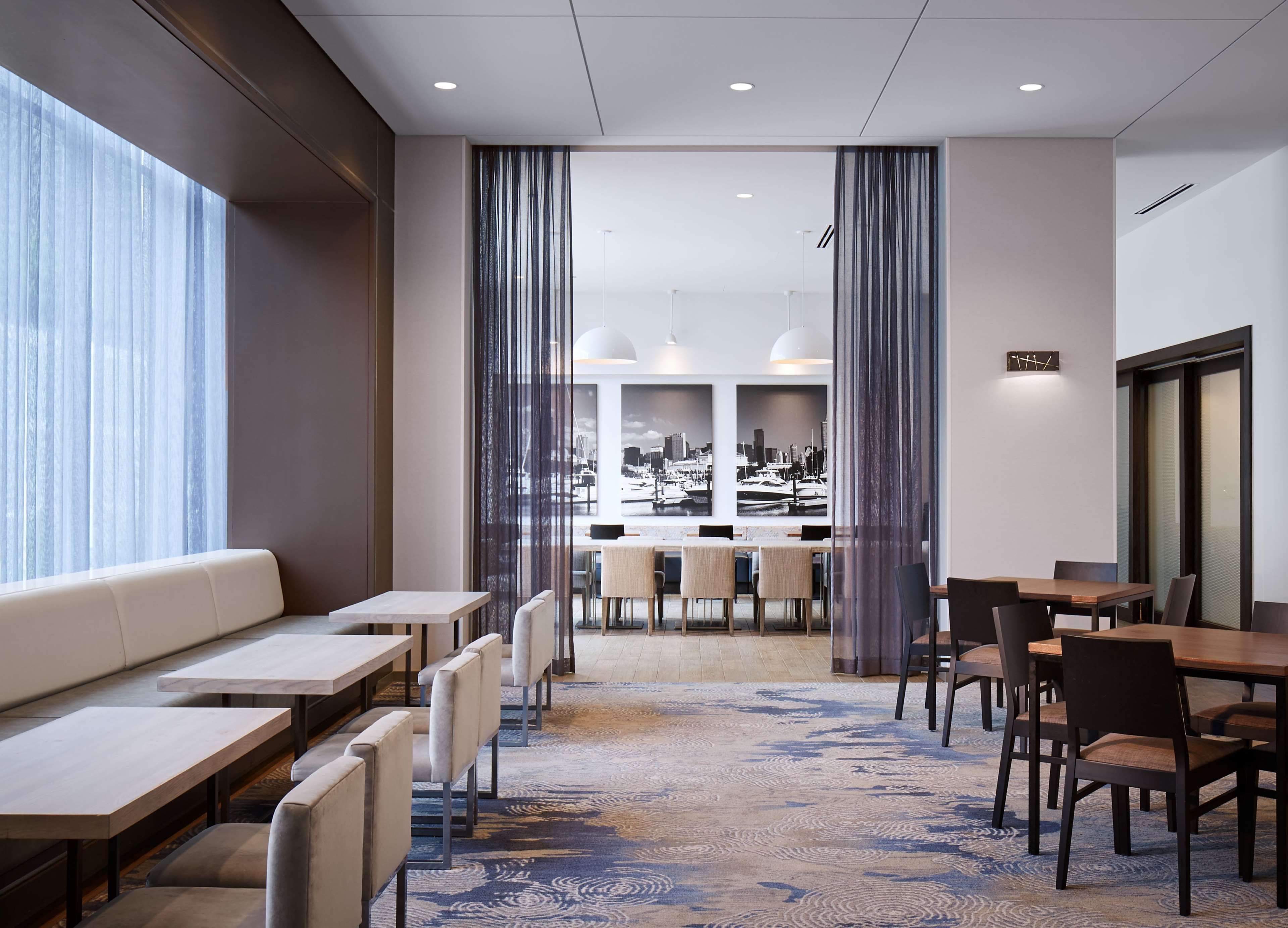 Un comedor de hotel moderno con una larga barra, diversas opciones de asientos y vistas a un puerto deportivo a través de amplios ventanales.