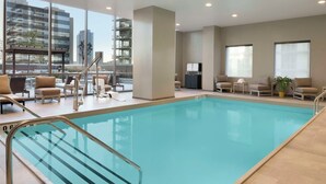 Una piscina interior con paredes de vidrio transparente, una escalera metálica y sillones beige para relajarse.