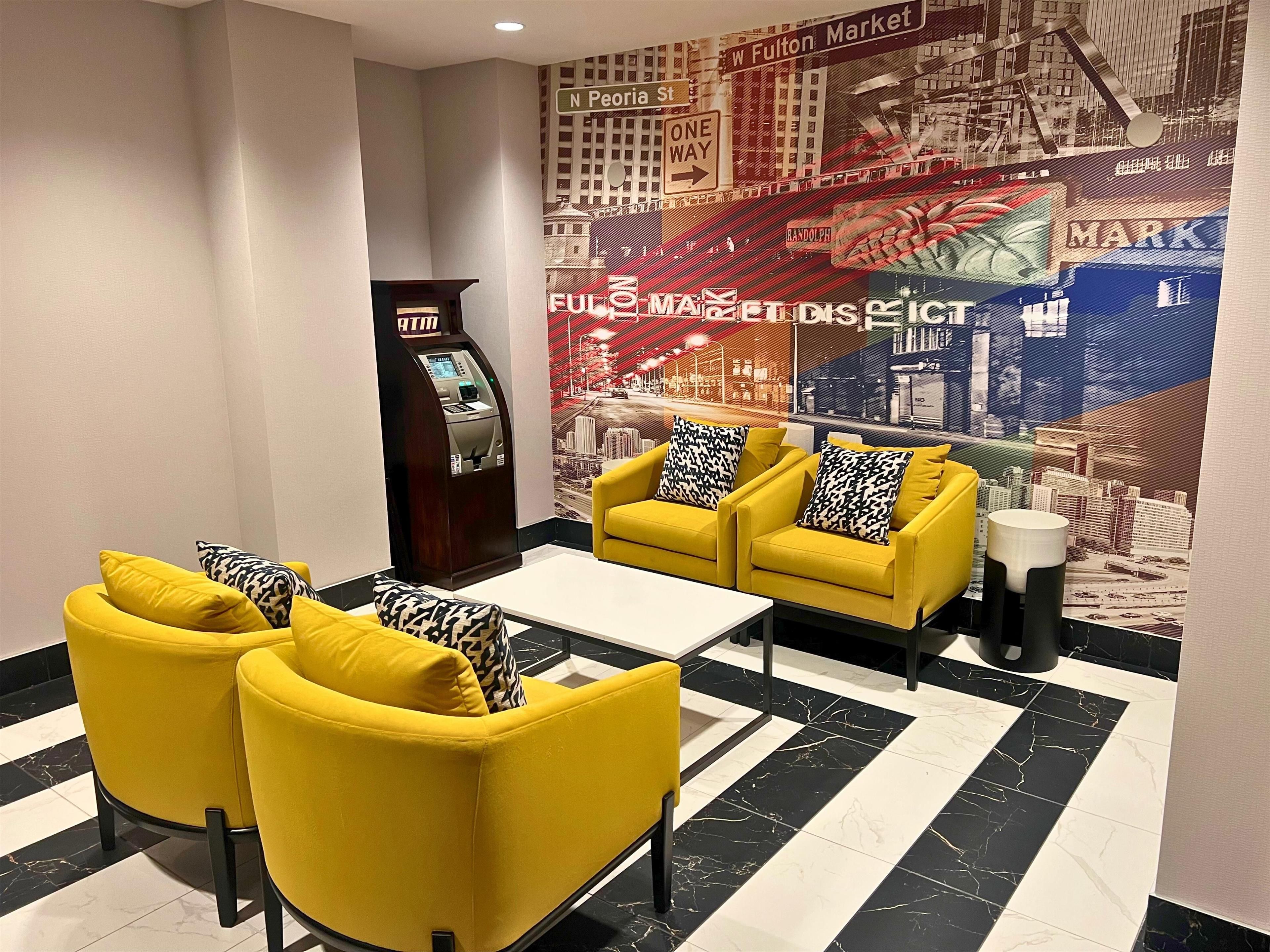 A lobby with yellow armchairs, a white coffee table, a cash machine, and a large cityscape mural on the wall.