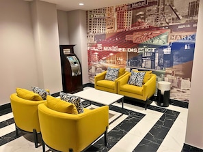 Un vestíbulo con sillones amarillos, una mesa de centro blanca, un cajero automático y un mural grande con vistas a la ciudad.