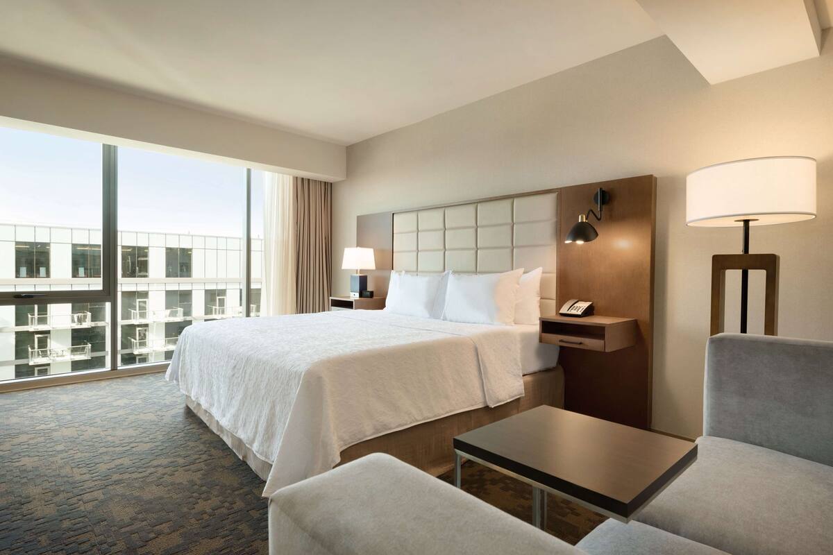 A hotel room with a large bed, a desk, a chair, and a view of a building through large windows.