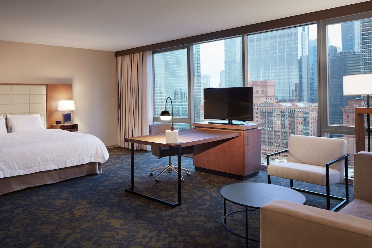 A hotel room with a bed, desk, chair, TV, and large windows with a city view.