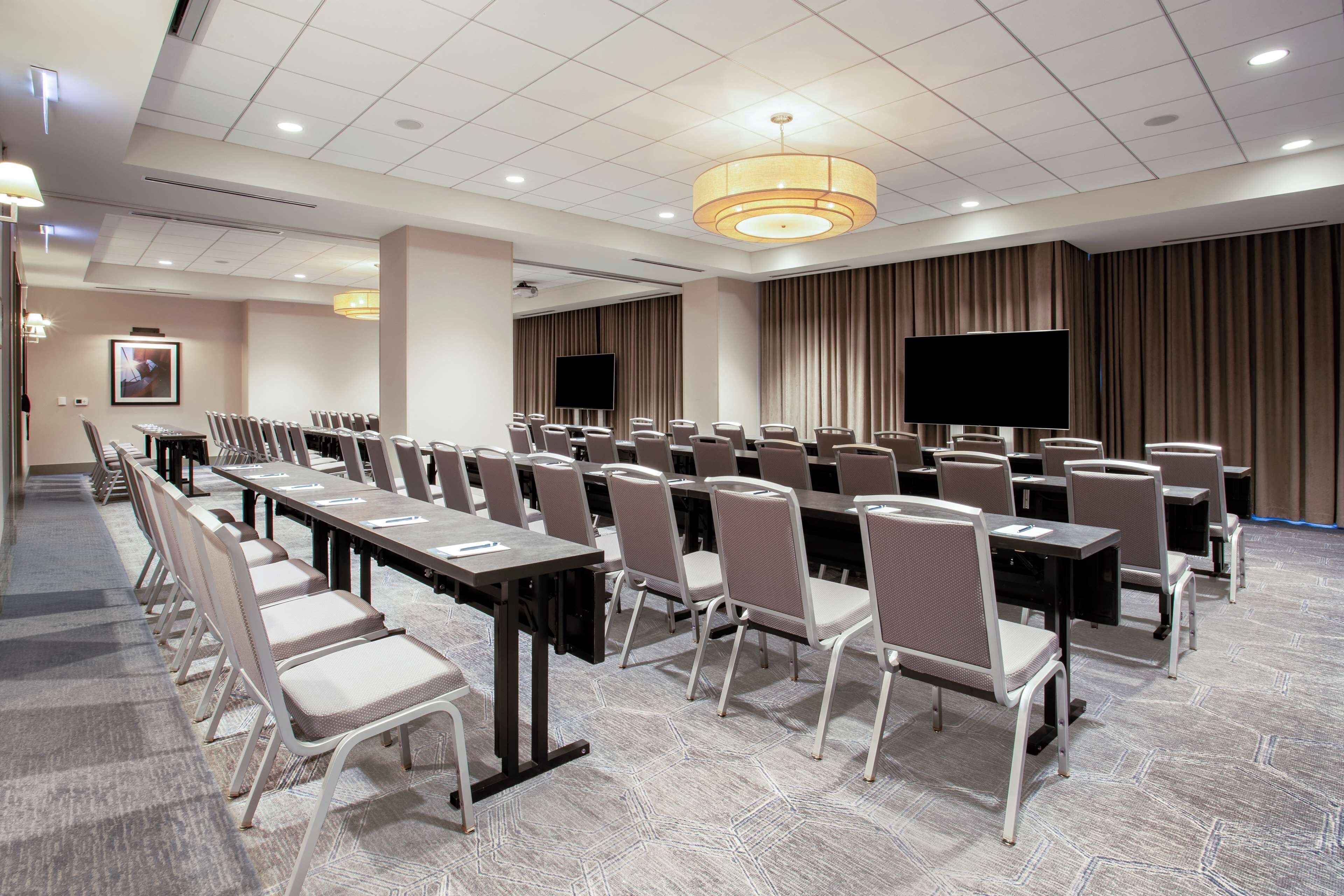 A conference room with rows of tables and chairs, a large flat-screen TV, and a ceiling-mounted light fixture.