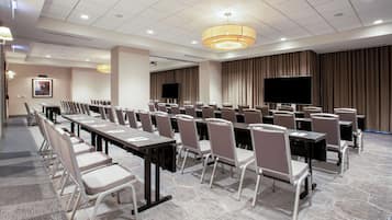 A conference room with rows of tables and chairs, a large flat-screen TV, and a ceiling-mounted light fixture.