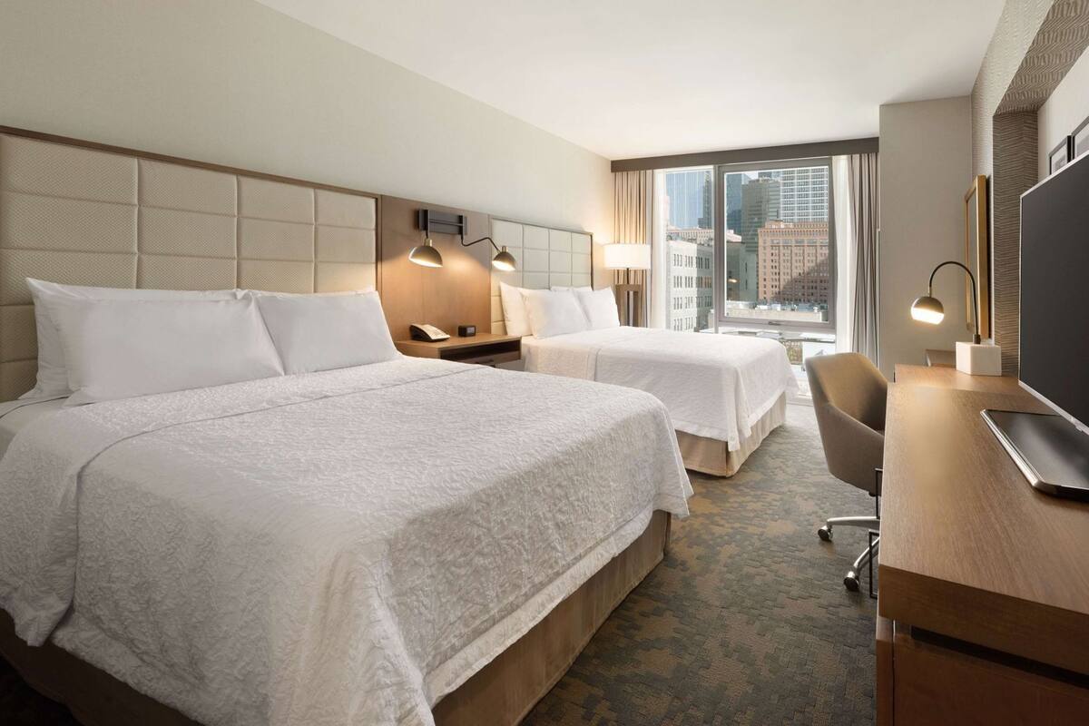 A hotel room with two beds, a desk, a chair, a TV, and a window with a city view.