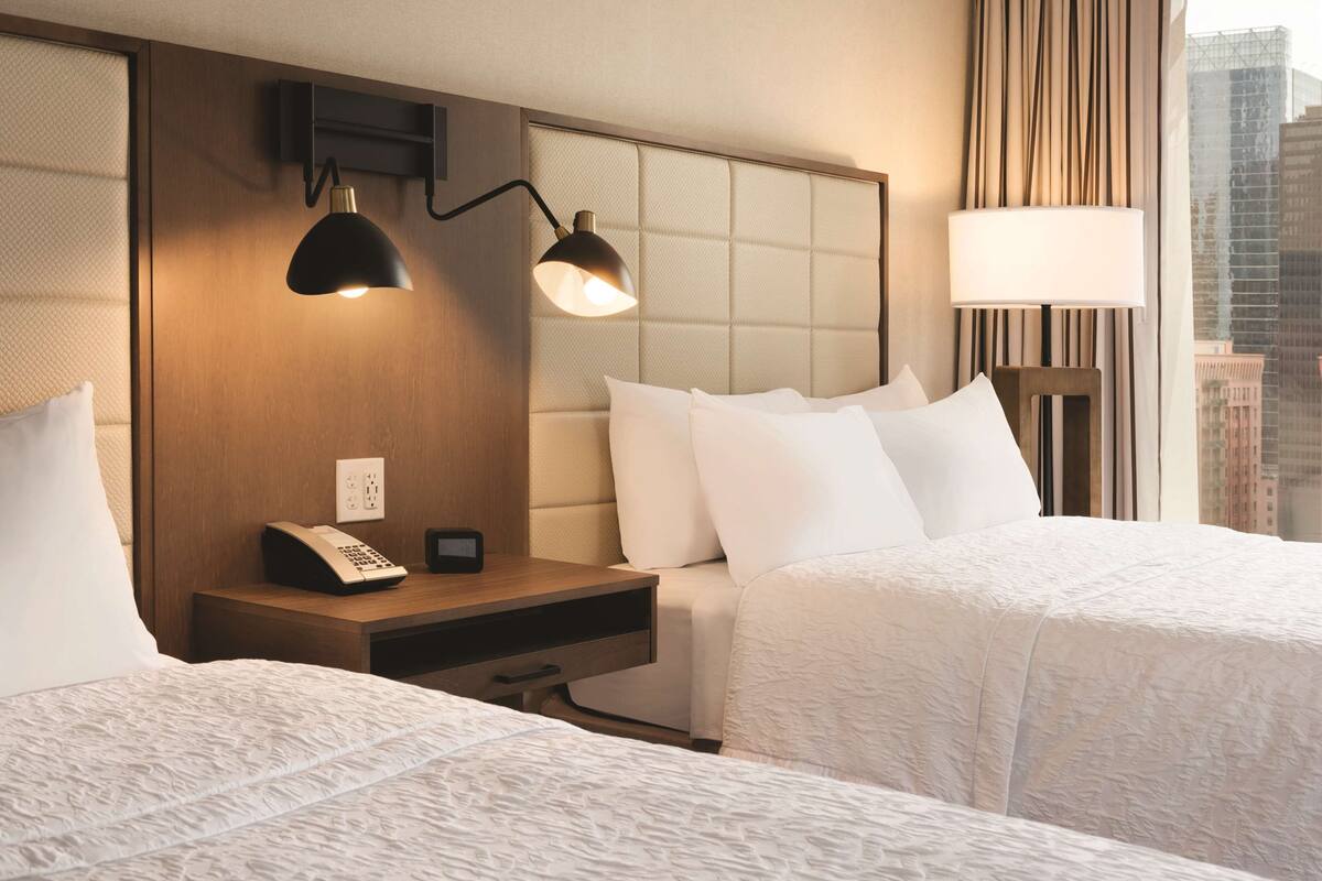 A hotel room with two beds, a nightstand with a telephone, a wall-mounted lamp, and a window with a view of buildings.