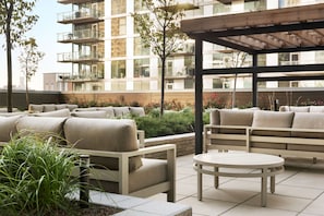 Un patio al aire libre con sofás color beige y una mesa redonda, rodeado de follaje y un edificio moderno con balcones al fondo.