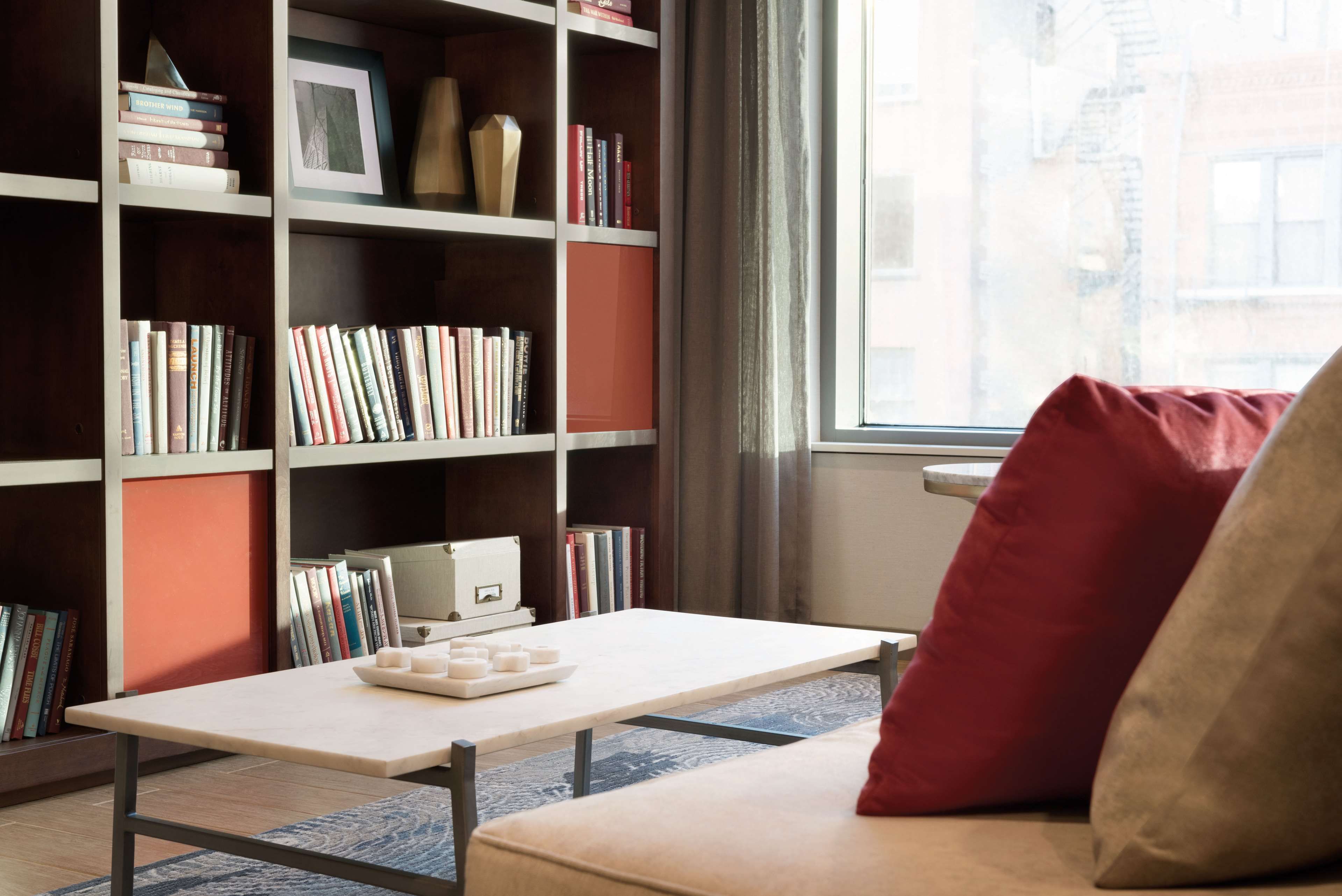 Una habitación con una estantería, una mesa llena de libros, un ventanal y un sofá con un cojín rojo.