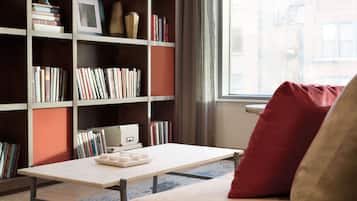 Una habitación con una estantería, una mesa llena de libros, un ventanal y un sofá con un cojín rojo.