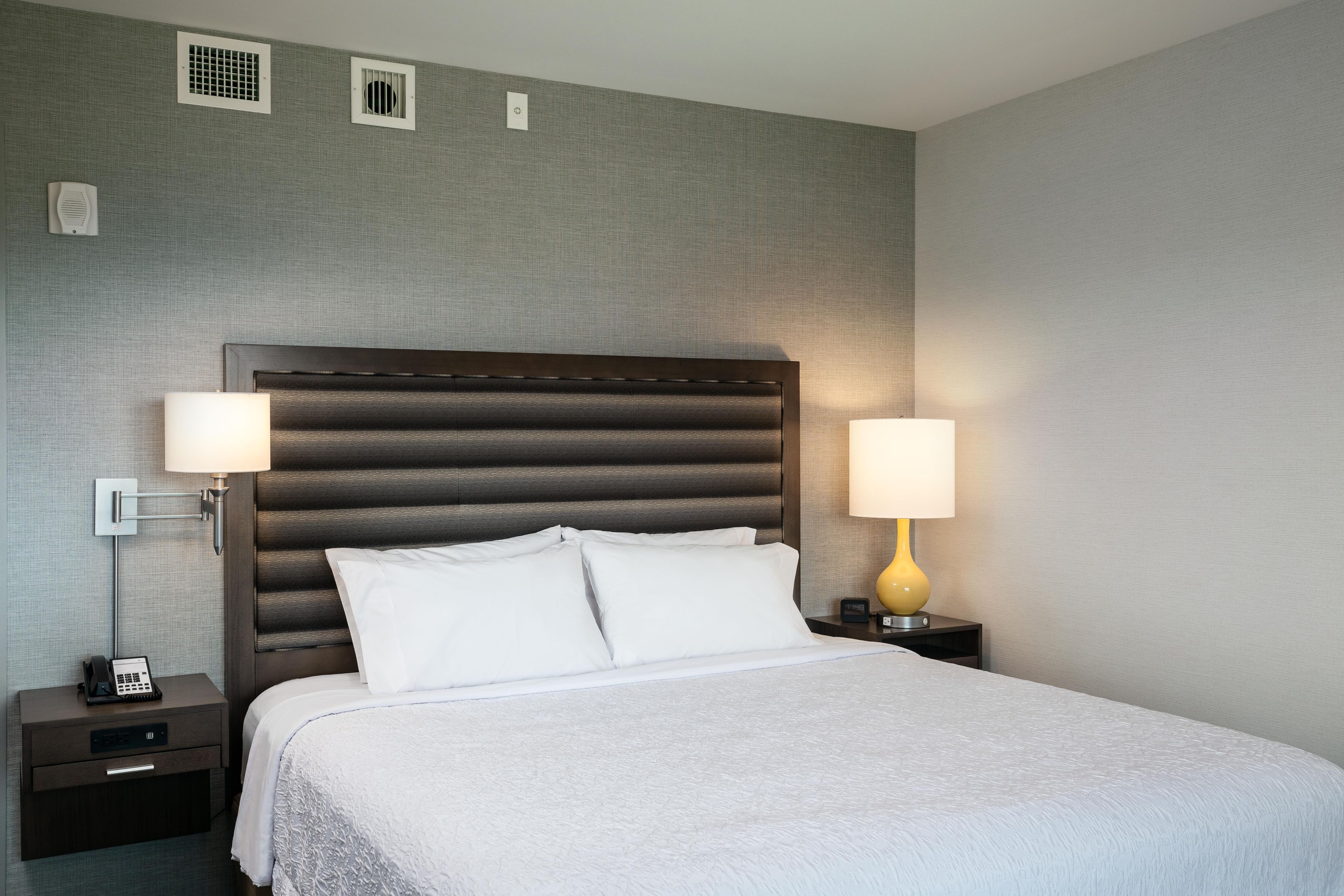 A hotel room with a large bed, two bedside tables, a nightstand, and a wall-mounted heater.