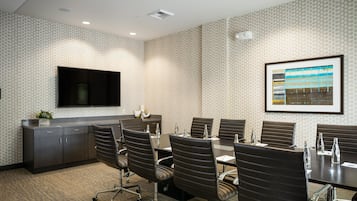 A conference room with a long table, multiple chairs, a flat-screen TV mounted on the wall, and a framed artwork above the table.