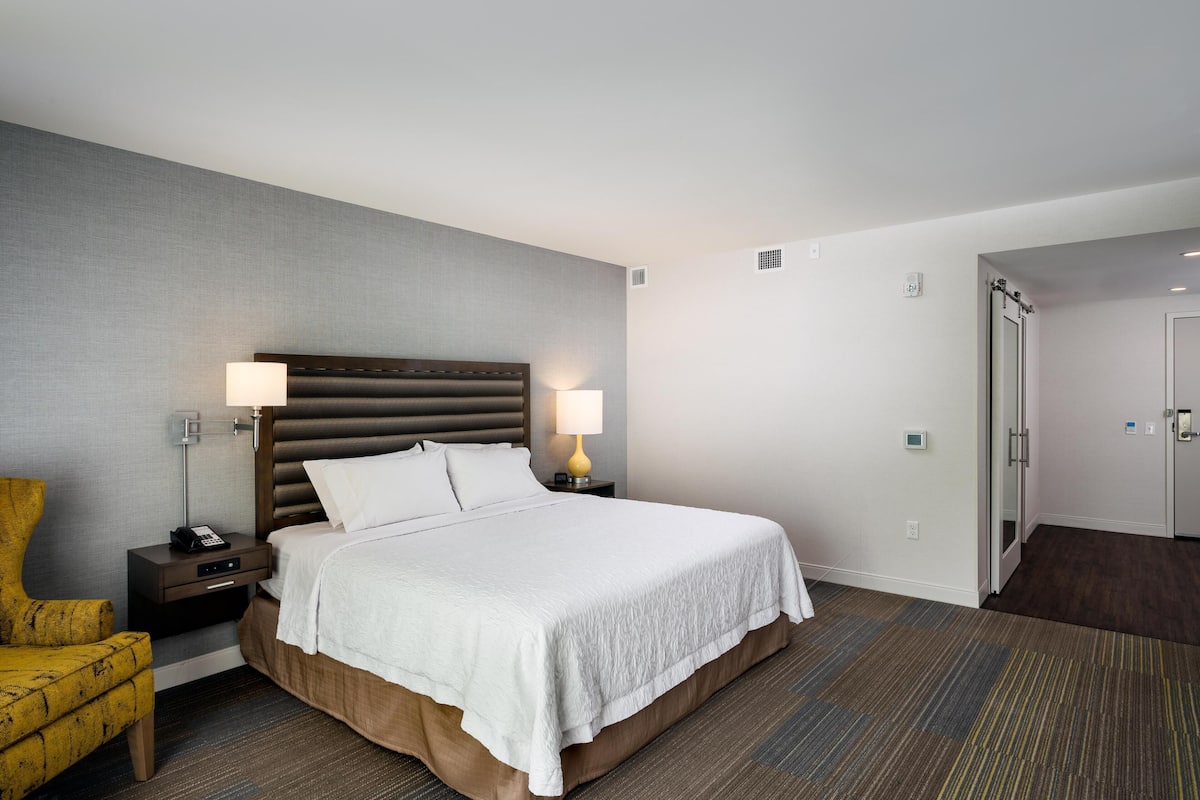 A hotel room with a large bed, two bedside lamps, a nightstand, a chair, and a door leading to another room.