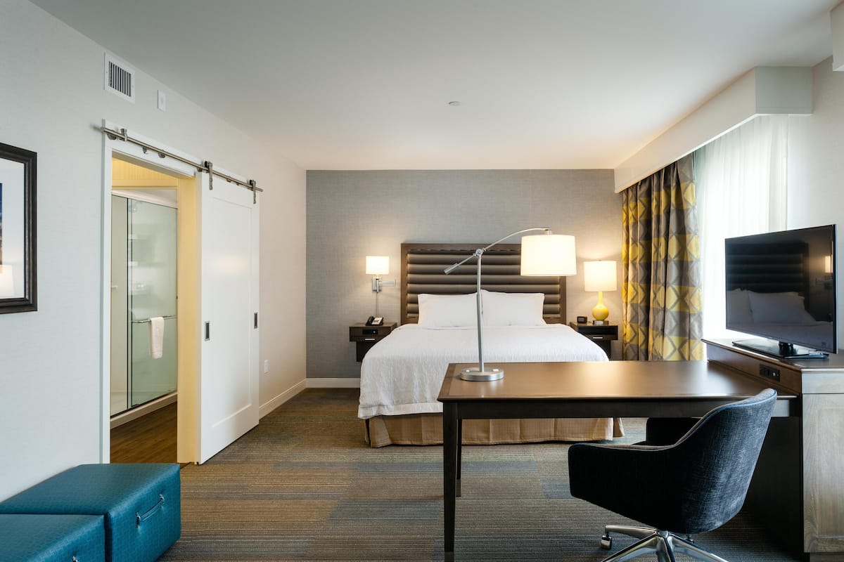 A hotel room with a bed, bedside tables, a desk, a chair, a television, and a bathroom with a glass door.