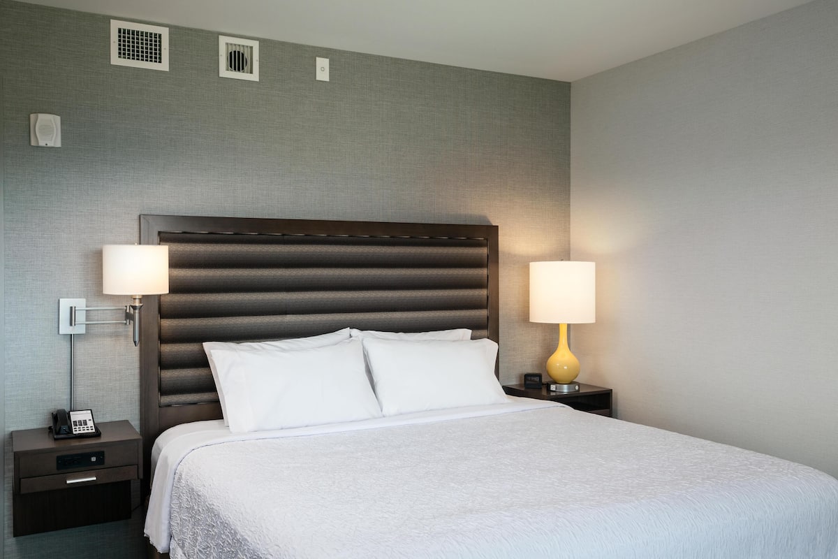 A hotel room with a large bed, two bedside lamps, a nightstand with a phone, and a wall-mounted vent.