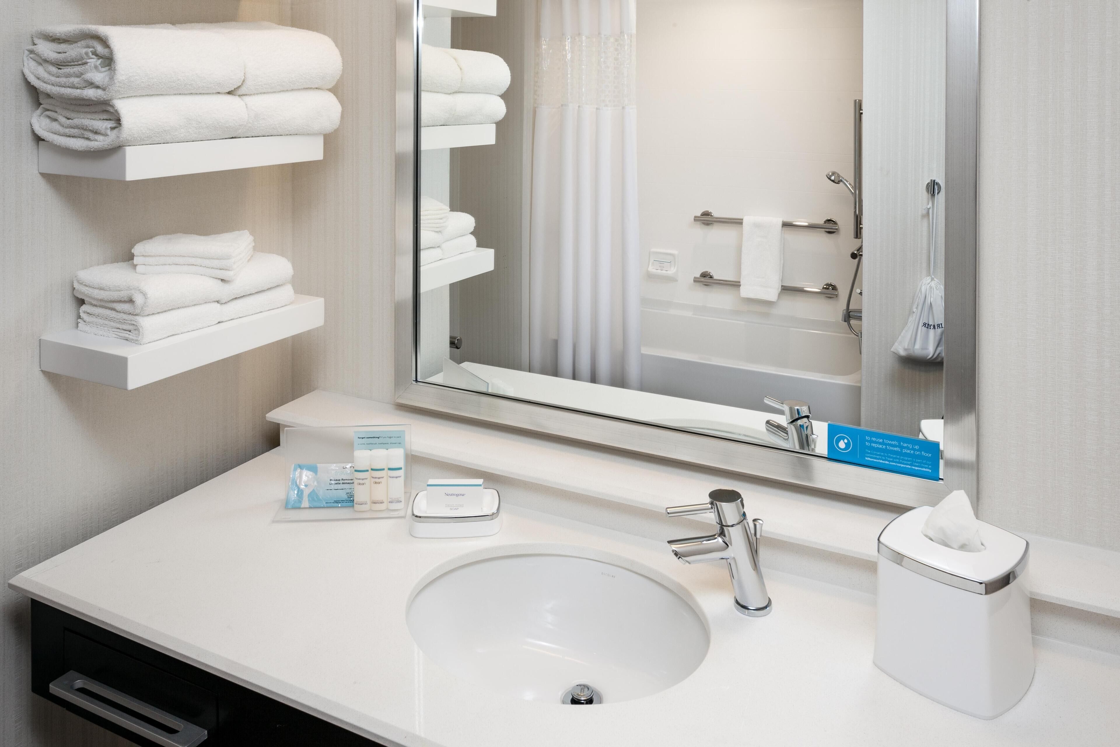 Une salle de bain d’hôtel avec un lavabo blanc, un miroir, un porte-serviettes et un distributeur de serviettes pour les mains.