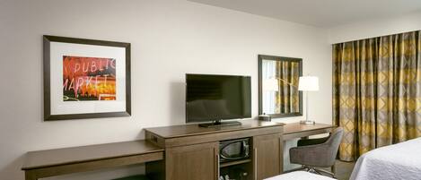 A hotel room with a bed, a desk with a TV, a lamp, a microwave, and a framed picture on the wall.
