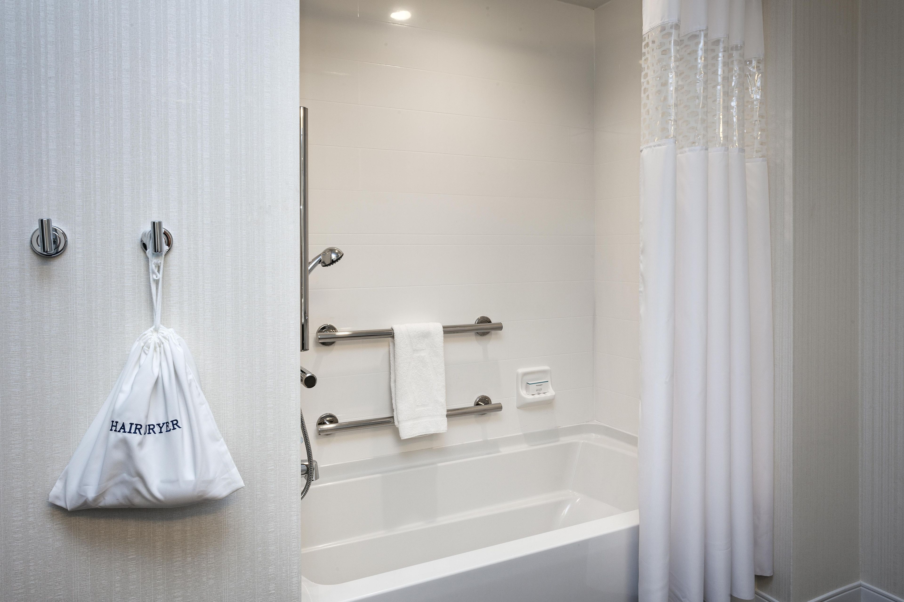 A bathroom with a bathtub, a towel rack with a towel, a soap dispenser, and a white bag hanging on the wall.