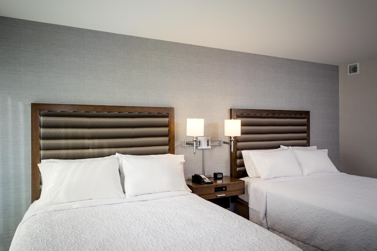 A hotel room with two beds, a nightstand with a lamp, and a wall-mounted light fixture.