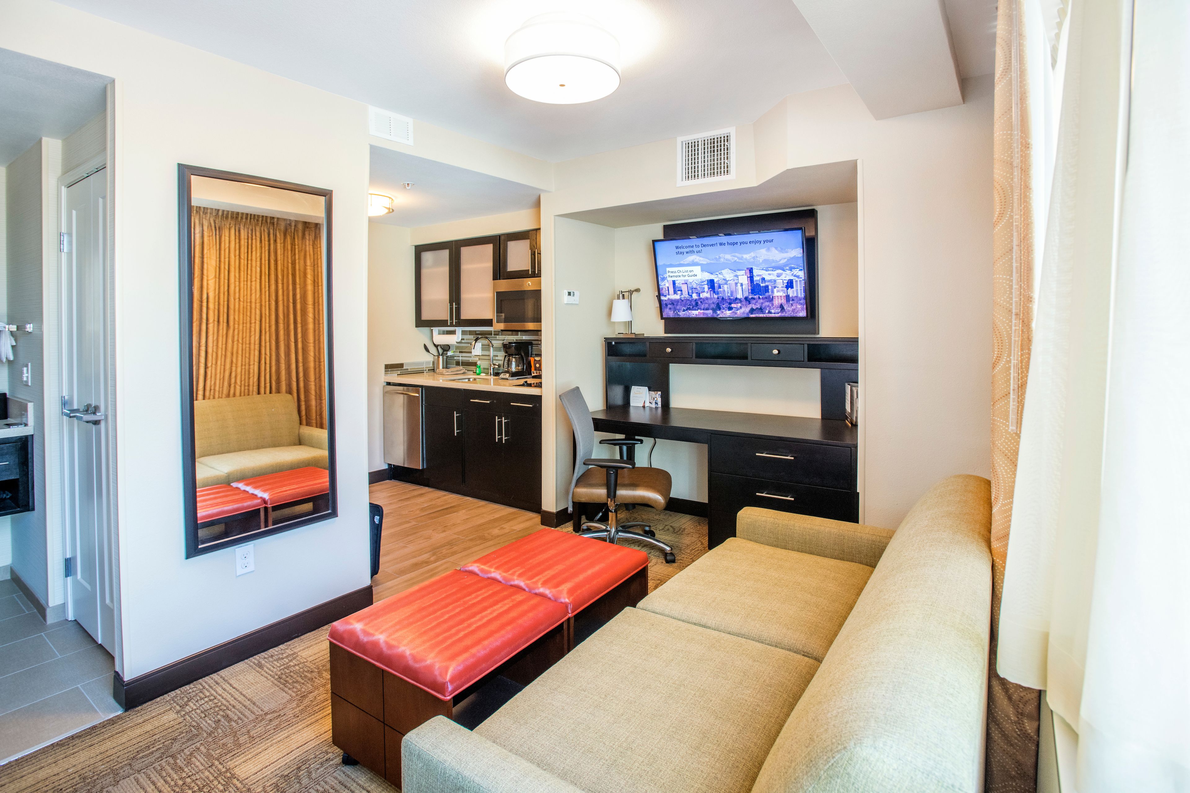 A hotel room with a sofa, a small table, a TV, a mirror, and a kitchenette.