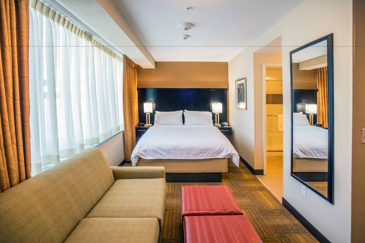 A hotel room with a large bed, two bedside tables with lamps, a sofa, and a red bench.