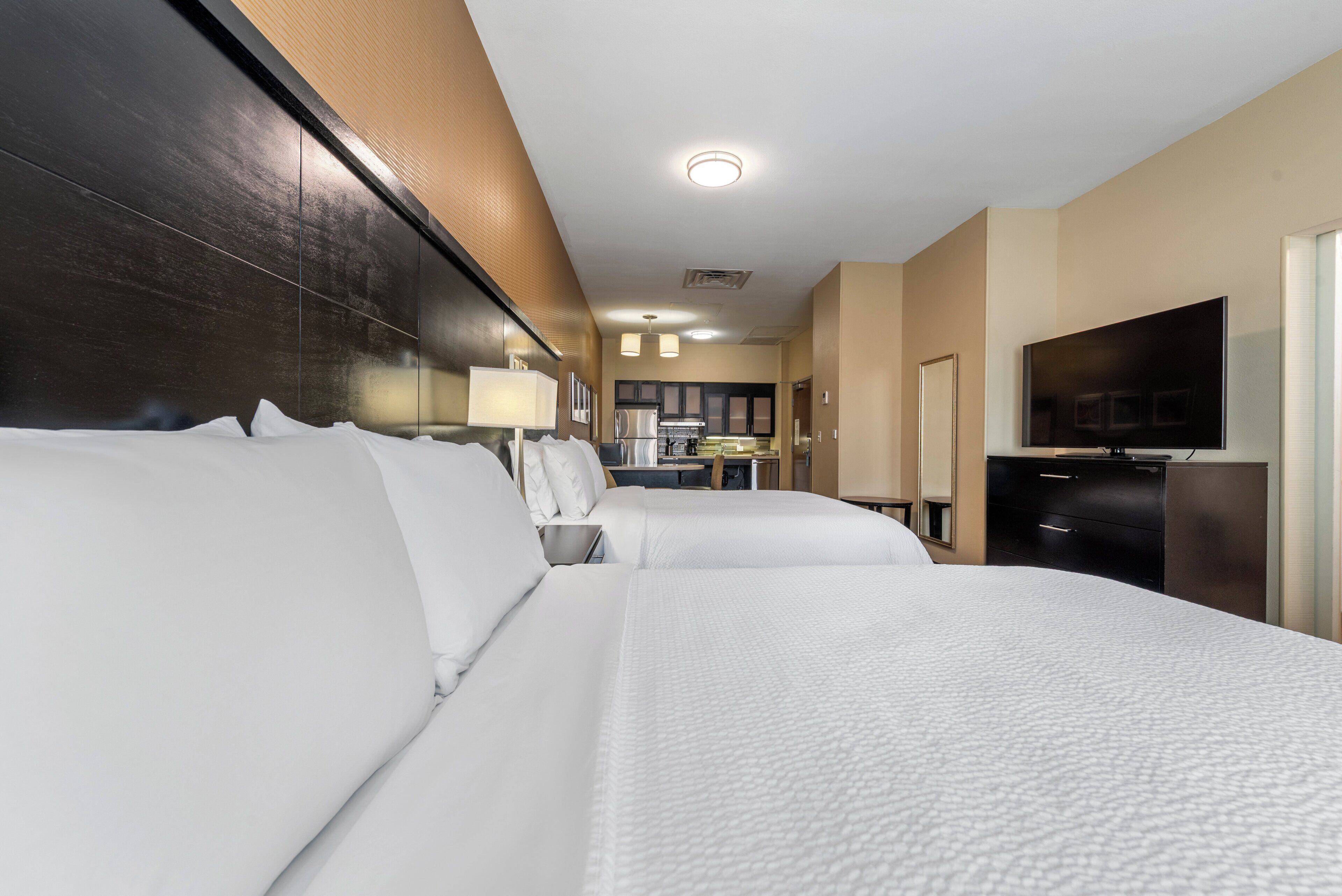 Ein Hotelzimmer mit zwei Betten, einem Fernseher, einem Schreibtisch und einer kleinen Küche im Hintergrund.