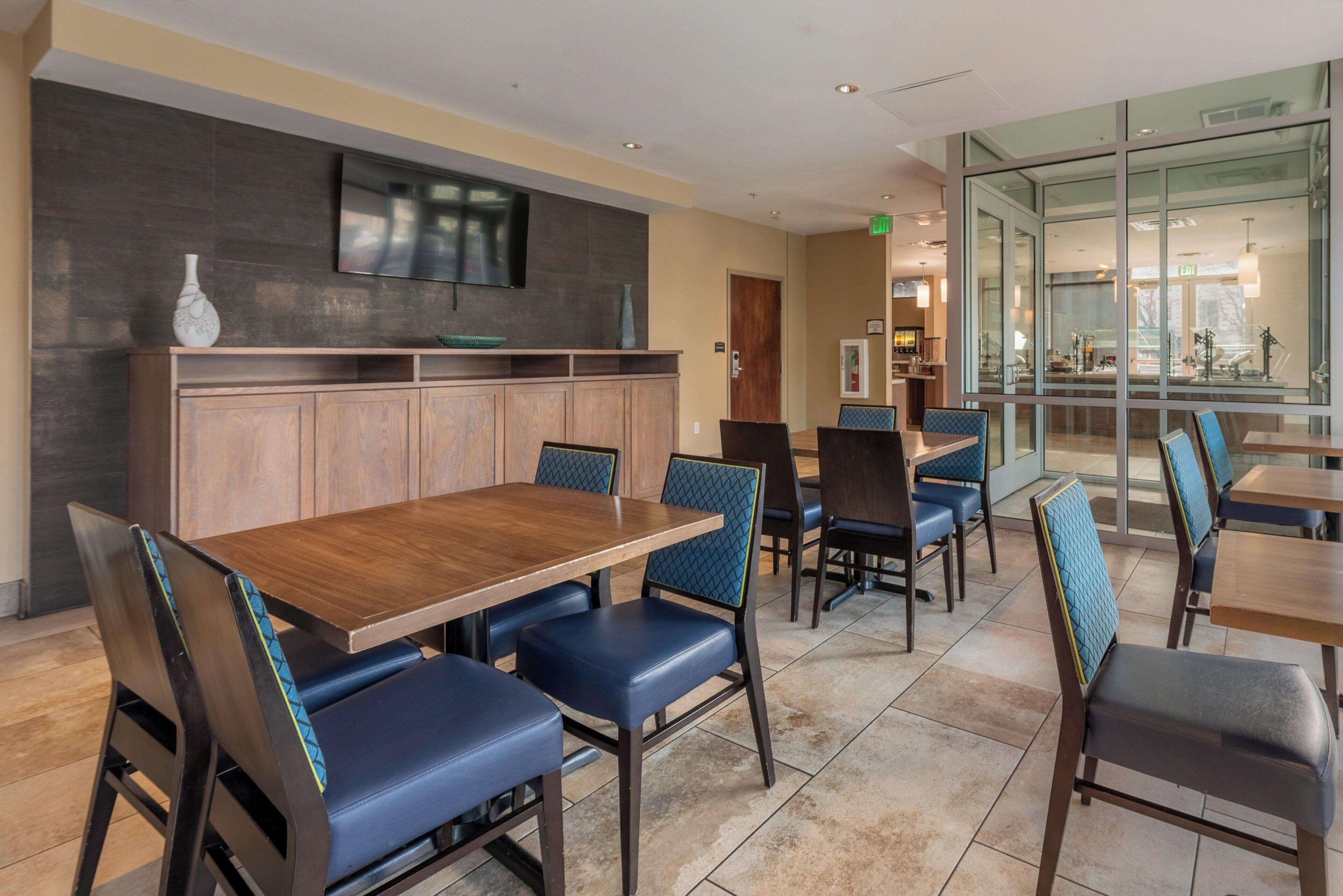 A dining area with a wooden table and blue upholstered chairs, a flat-screen TV mounted on the wall, and a glass partition separating the dining area from another space.